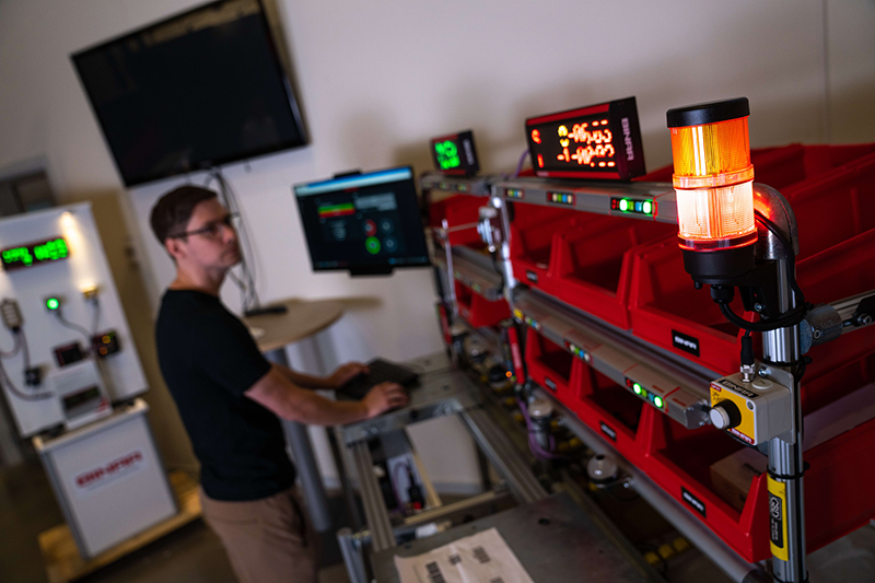 A warehouse employee is interacting with a pick2light system, standing by a workstation with illuminated digital displays and an orange light signal indicating activity or a notification.